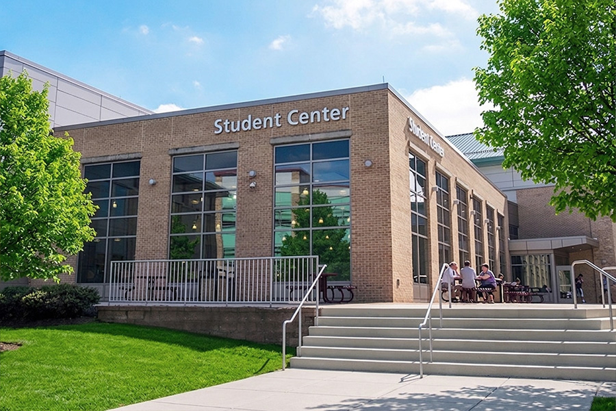 Exterior view of the Frederick Community College Student Center building, with large glass windows, a staircase, and students seated at outdoor tables on a sunny day.