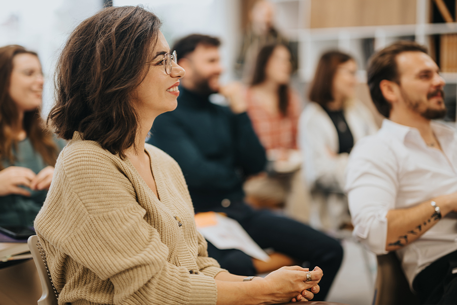 A smiling woman wearing glasses and a tan cardigan sits attentively in an orientation or classroom session, with other engaged adult participants visible in the background.