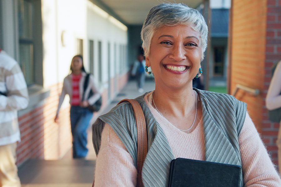 A smiling older adult woman with short gray hair holds a notebook and carries a shoulder bag while standing in an outdoor campus corridor, with other students visible in the background.