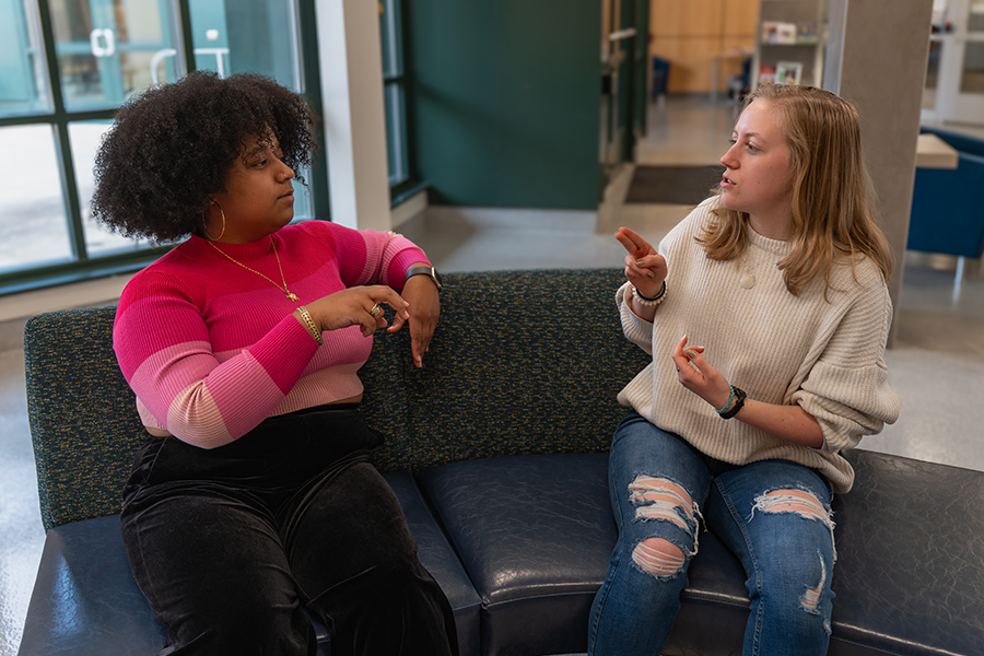 Two students sit on a couch in a campus lounge, talking and gesturing with their hands.