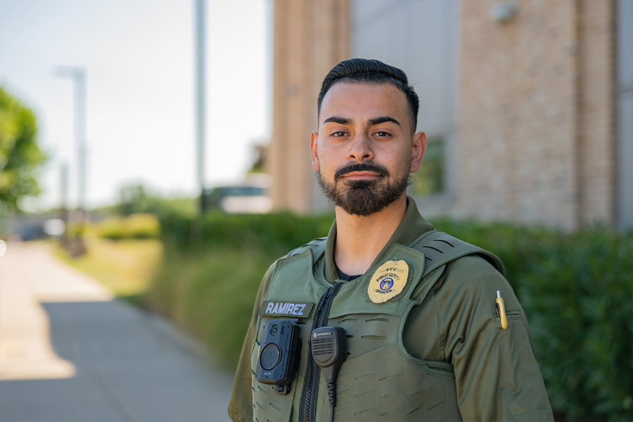 Close-up of a bearded public safety officer named Ramirez in an olive green uniform and tactical vest, featuring a badge and body camera, outdoors.