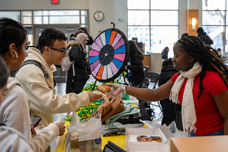 At a vibrant club fair, a student in a red shirt and white scarf smiles while handing a donut to a fellow student across a table. The table is decorated with yellow and green tinsel and features a colorful prize wheel in the background. Other students are seen mingling and visiting booths in the brightly lit student center.