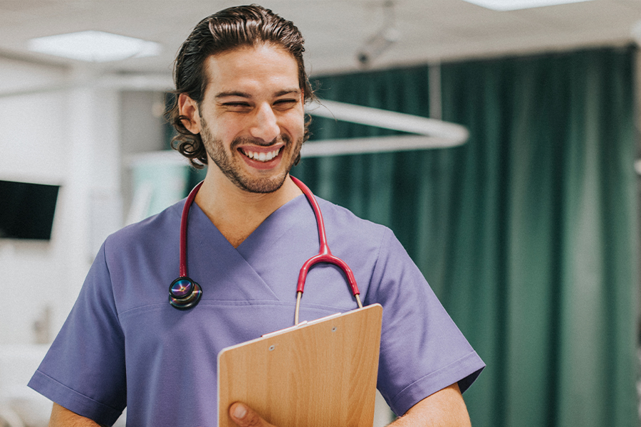 A smiling male healthcare professional in purple scrubs with a red stethoscope holds a clipboard in a hospital room with green privacy curtains visible in the background.