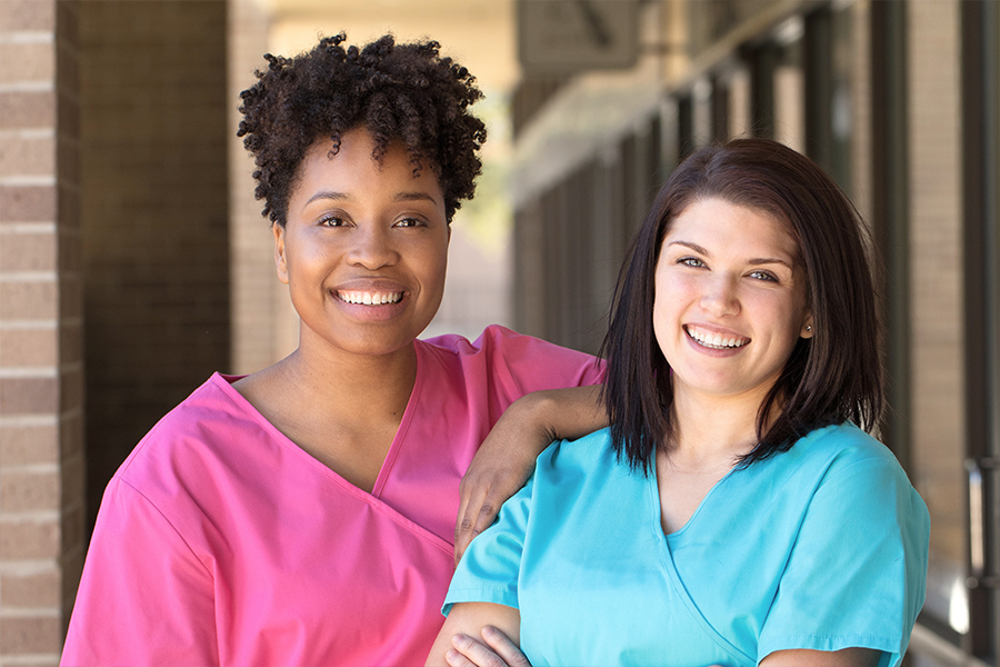 Two smiling healthcare students in scrubs standing together outside a building, one in pink scrubs and one in teal scrubs.