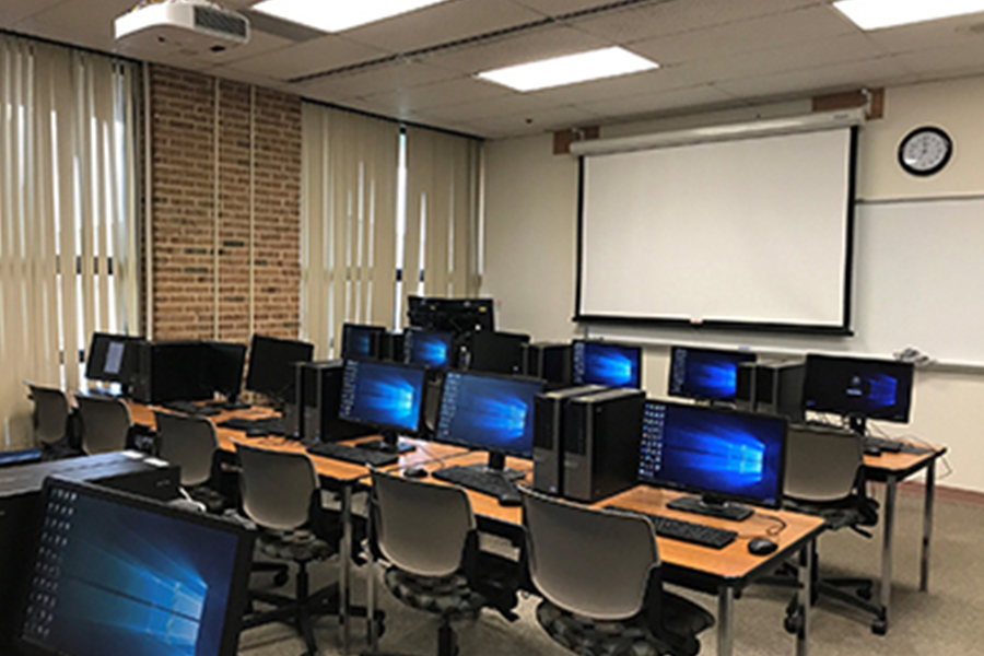 A classroom featuring rows of desktop computers, black office chairs, and a large projector screen at the front of the room.