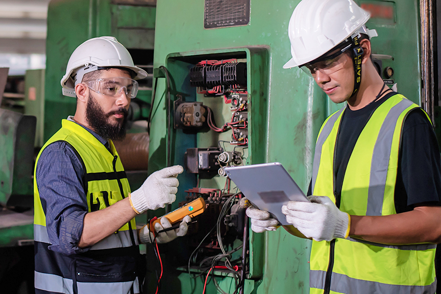 Two electricians wearing white hard hats, safety glasses, and high-visibility vests review a tablet together while working near an industrial electrical panel.
