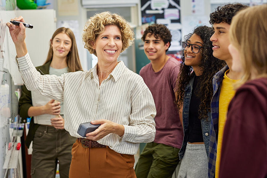 A smiling instructor writes on a whiteboard while a diverse group of five students gathers around her attentively in a busy classroom.