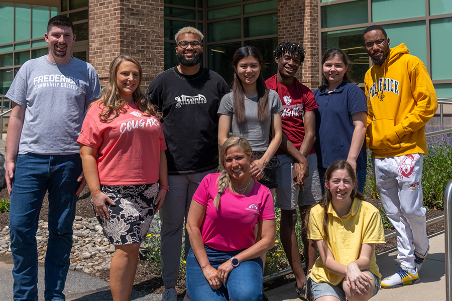 A group of nine students and staff smile together outside an FCC campus building on a sunny day, several wearing Frederick Community College apparel.