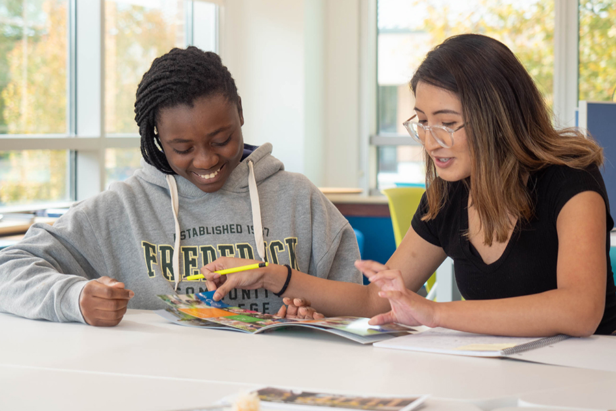 Two students sit together at a white table in a brightly lit room. One student, wearing a grey Frederick Community College hoodie, looks on and smiles as the other student points to a colorful brochure and explains something.