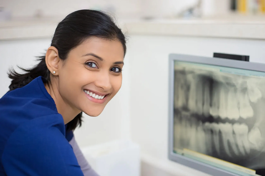 A smiling dental professional in a blue uniform turns toward the camera while a dental X-ray image is displayed on a monitor behind her.