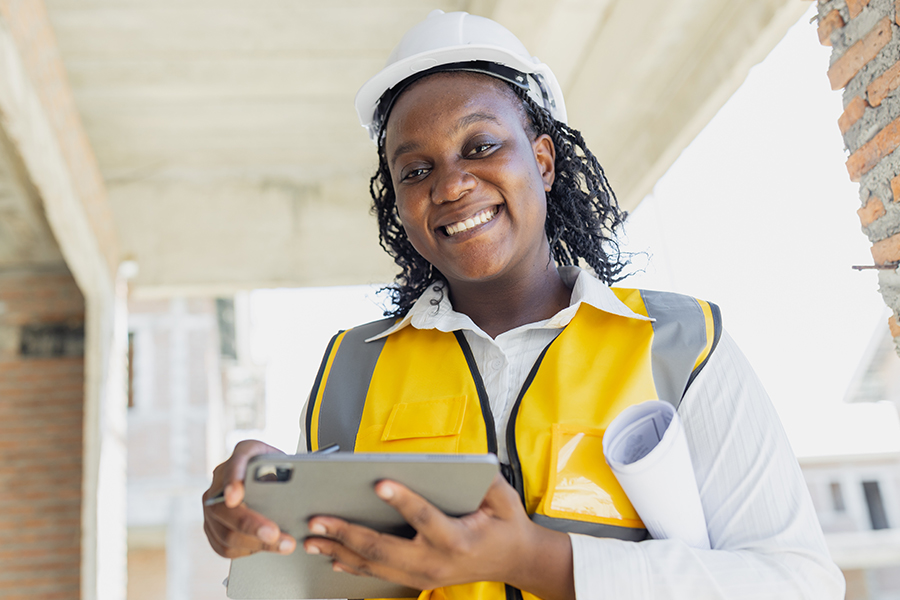 A smiling woman wearing a white hard hat and yellow safety vest holds a tablet and rolled blueprints while standing on the porch of a building under construction.