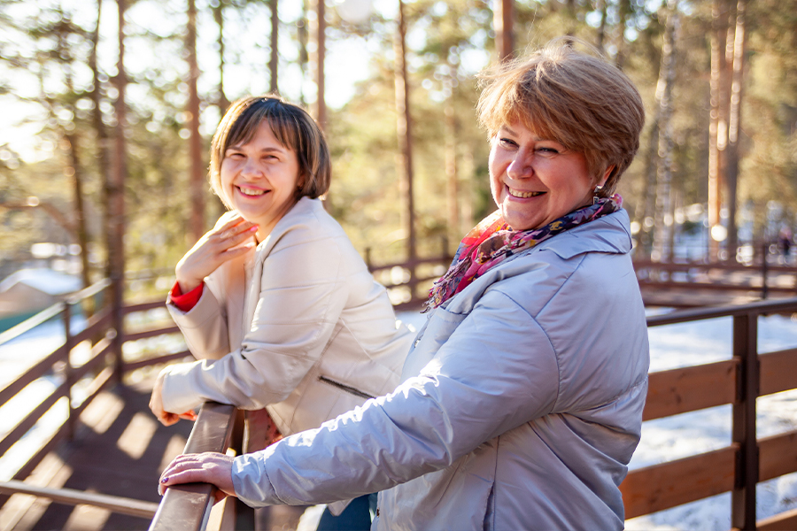 Two smiling women lean on a wooden railing together outdoors, surrounded by sunlit trees in winter.