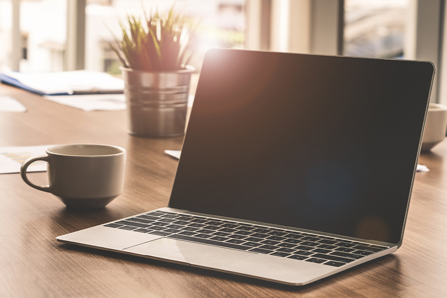 An open laptop sits on a wooden desk next to a coffee mug and a potted plant in a bright, sunlit office.