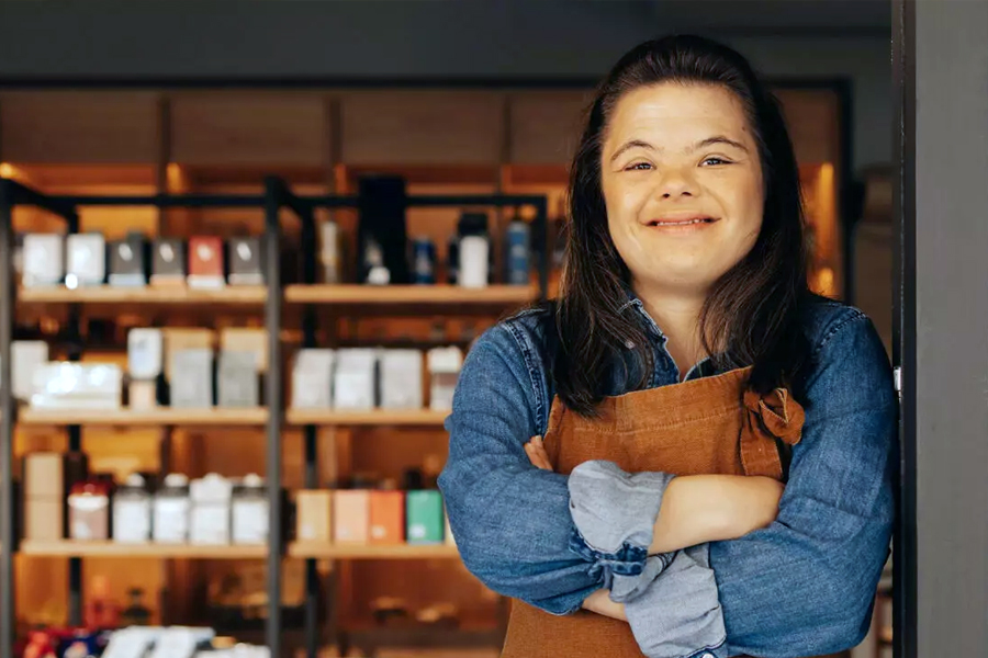 A smiling woman wearing a denim shirt and brown apron stands with arms crossed in a shop, with shelves of products visible behind her.