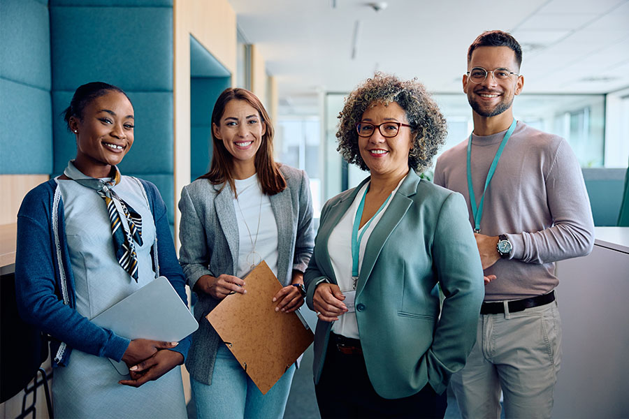 Four smiling professional adults stand together in a modern office setting, with one holding a laptop and another holding a clipboard. One woman wears a teal blazer and glasses, and a man in the background wears a lanyard.