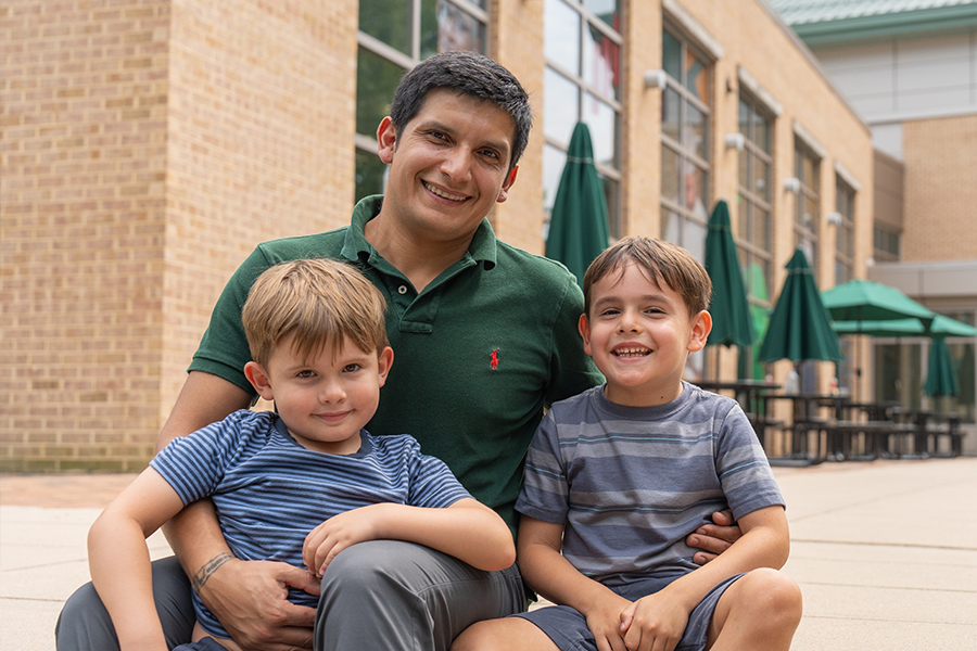 A smiling adult student in a green polo shirt sits outdoors on a campus patio, hugging his two young sons who are also smiling.
