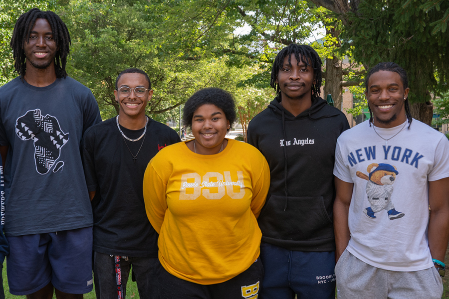 Five smiling students of color stand together in a row outdoors on a grassy campus area with trees in the background.