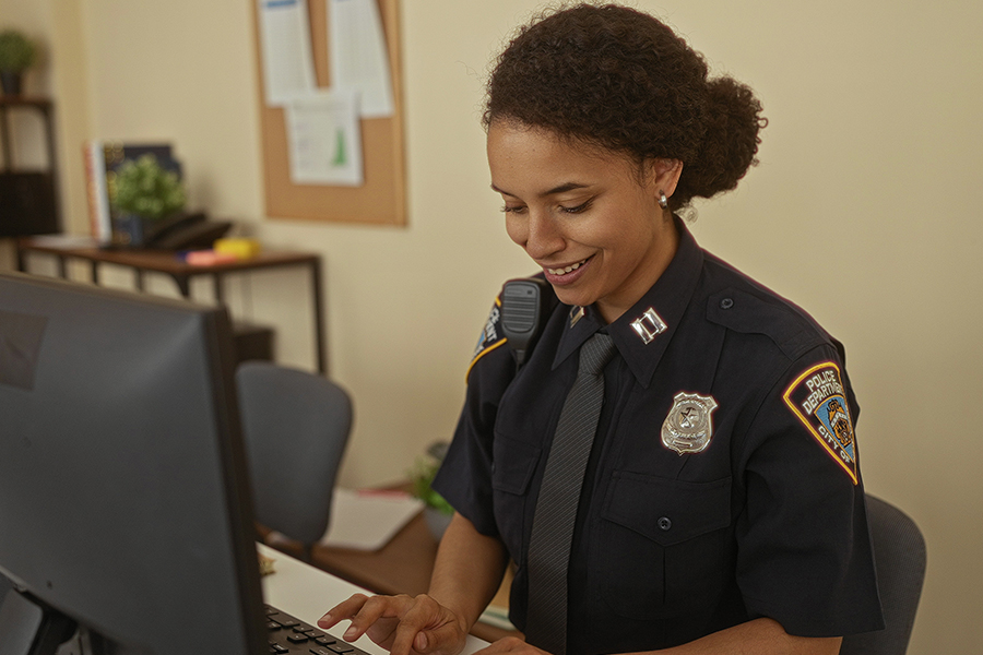 A female law enforcement officer in a navy blue uniform sits at a wooden desk, focused on a laptop while taking notes during an independent study session.