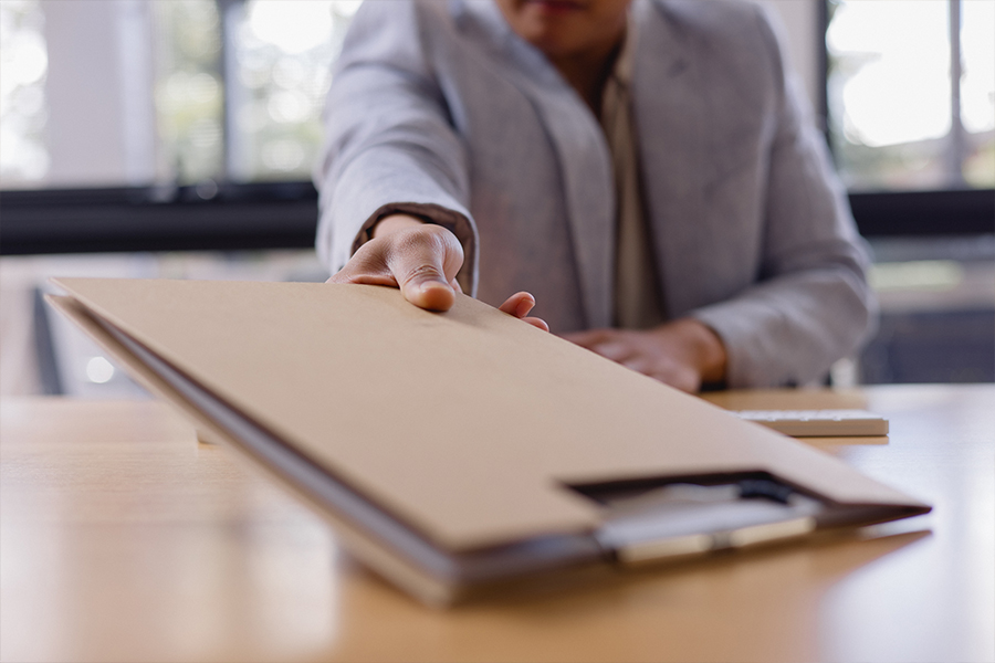 A first-person perspective shot of an office professional in a light grey blazer handing a tan folder or document across a wooden desk toward the viewer. The background is softly blurred, showing a bright window.