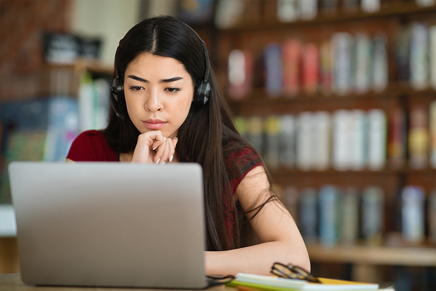A young woman with long dark hair wearing a red shirt and a headset sits at a desk, looking intently at a laptop screen. She has her hand on her chin in a thoughtful pose, with blurred library bookshelves in the background.