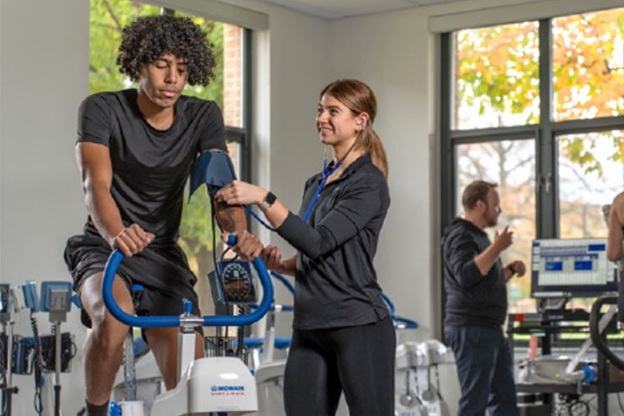 A student on a stationary exercise bike has his blood pressure monitored by a peer using a manual sphygmomanometer and stethoscope in a well-lit Human Performance Lab.