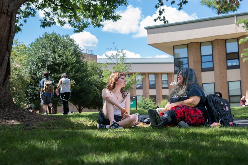 Two people are sitting on a grassy campus lawn in front of Frederick Community College's Braddock Hall, enjoying a clear day with a black backpack resting nearby.