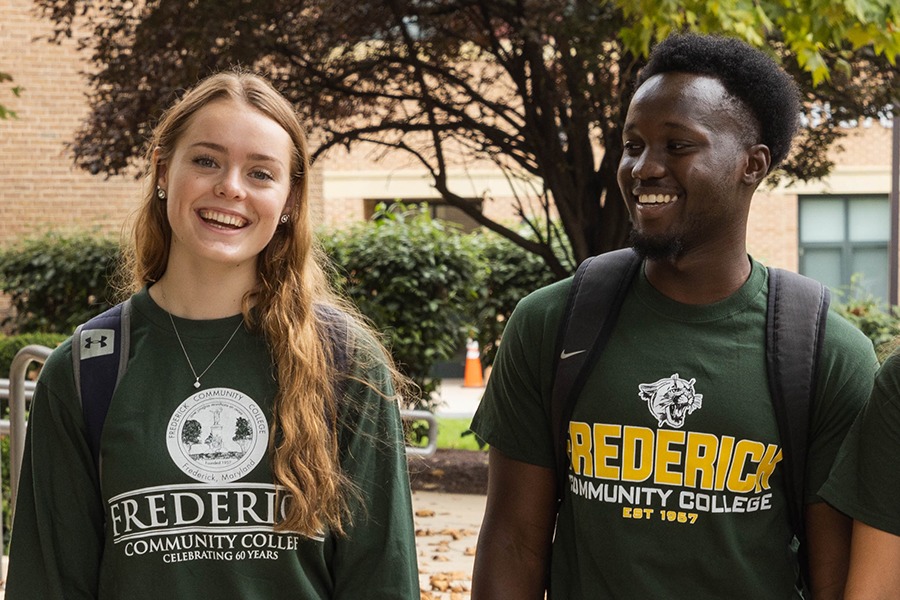 Two students in green shirts stand outside a building, smiling and engaged in conversation.