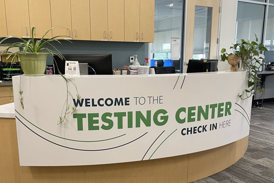 A bright, modern reception desk at the Frederick Community College Testing Center. The curved white desk features large green and navy blue text that reads, "WELCOME TO THE TESTING CENTER - CHECK IN HERE." Two potted green plants sit on either end of the counter, and computer monitors and office supplies are visible behind the desk in a professional, well-lit office setting.