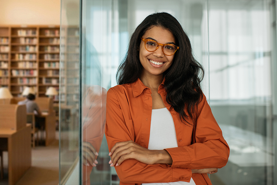 A smiling young woman in an orange shirt and glasses stands with arms crossed near the entrance of a library.