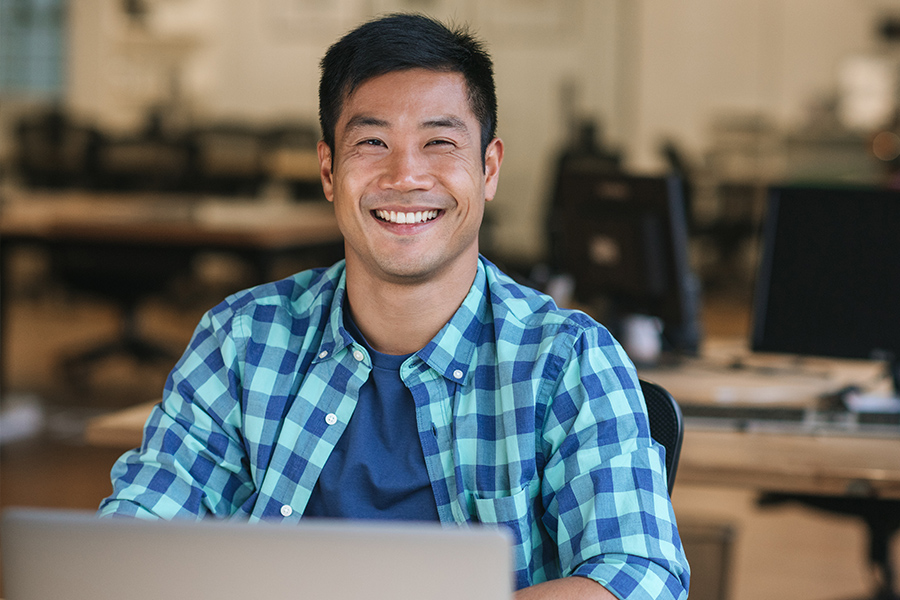 A smiling man in a blue checkered shirt sits at a laptop in an open office setting.