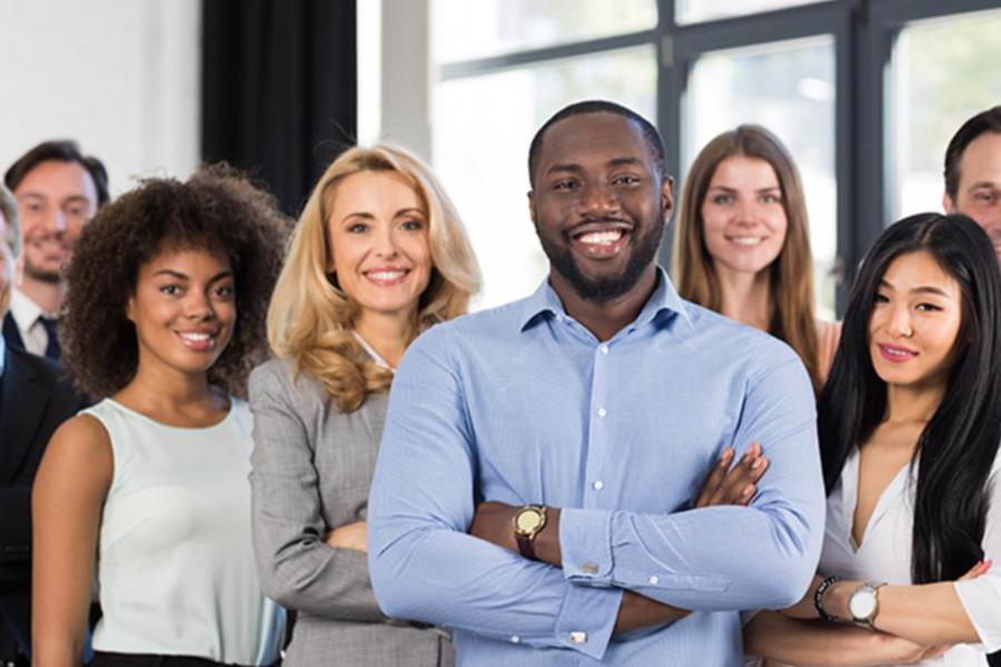 A diverse group of smiling business professionals standing together in an office, with a man in a light blue shirt crossing his arms in the foreground.