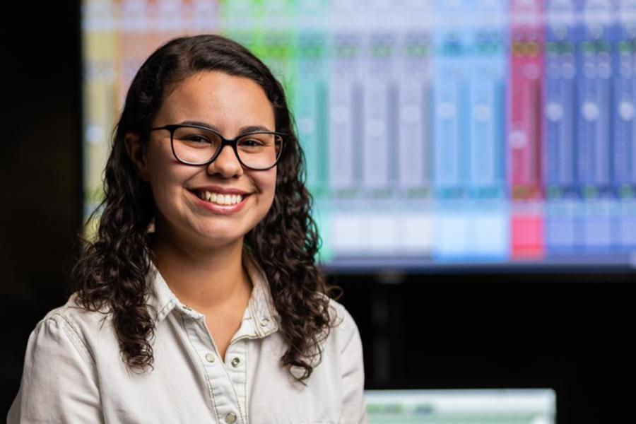 A smiling female student with curly dark hair and glasses in a recording studio, with a colorful digital audio workstation interface visible in the background.