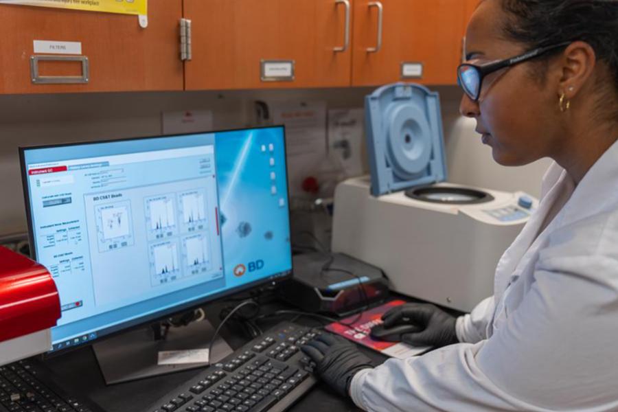 A woman in a lab coat and gloves reviews data on a computer monitor in a laboratory setting.