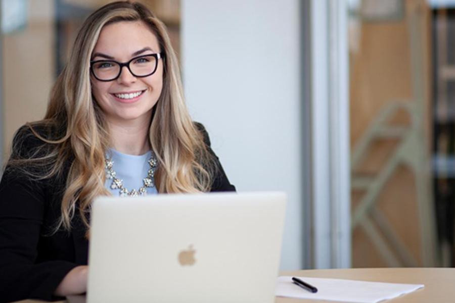 A smiling professional woman with blonde hair and glasses working at a laptop in an office setting.