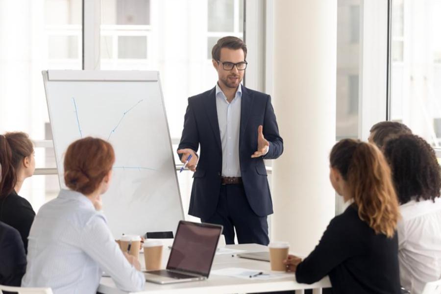 A male professional in a suit and glasses giving a presentation to a group in a bright, modern conference room.