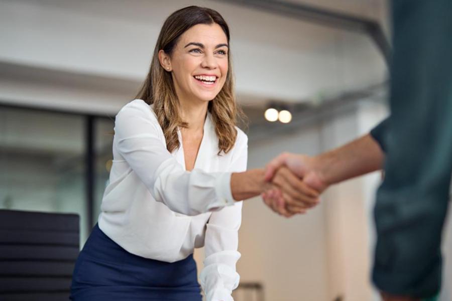 A professional woman in a white blouse smiles broadly while shaking hands with an unseen colleague in a bright, modern office setting.