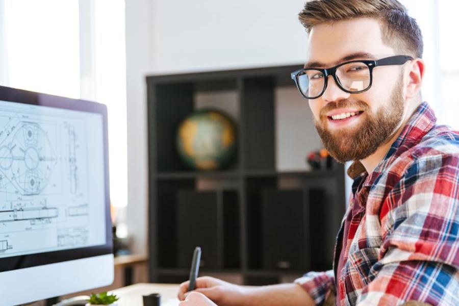 A smiling man with a beard and glasses sits at a desk with a computer displaying a technical blueprint.