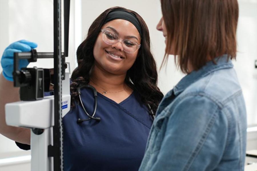 A healthcare professional in blue scrubs adjusts a balance beam scale while smiling at a patient.