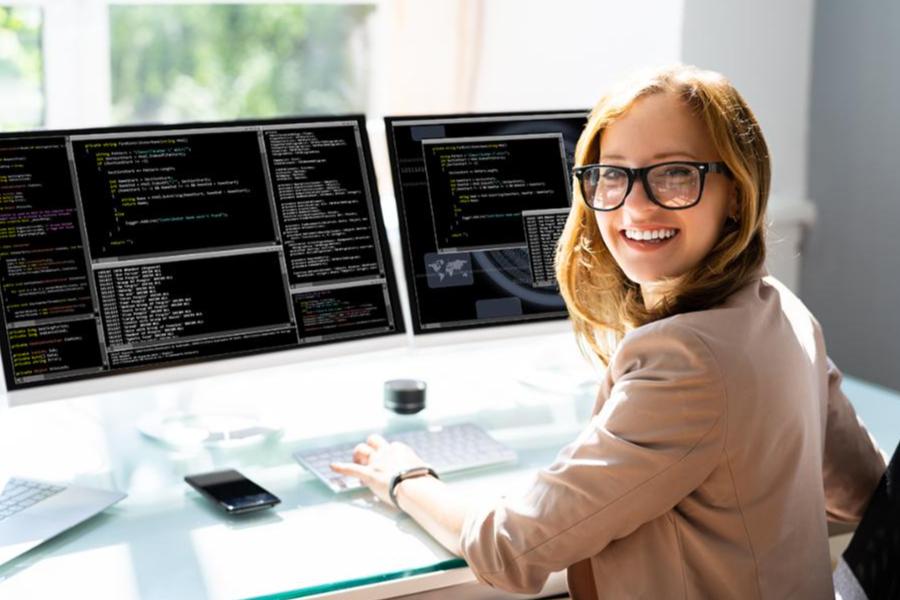 A smiling woman with glasses sits at a desk with two computer monitors displaying lines of code.