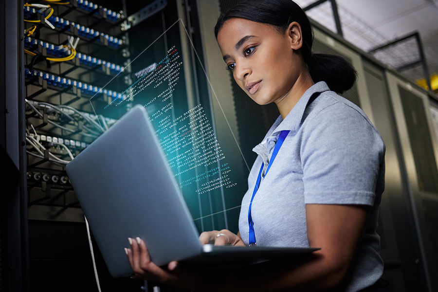 A female IT professional holds a laptop while working in a server room, with racks of network cables and equipment visible behind her.