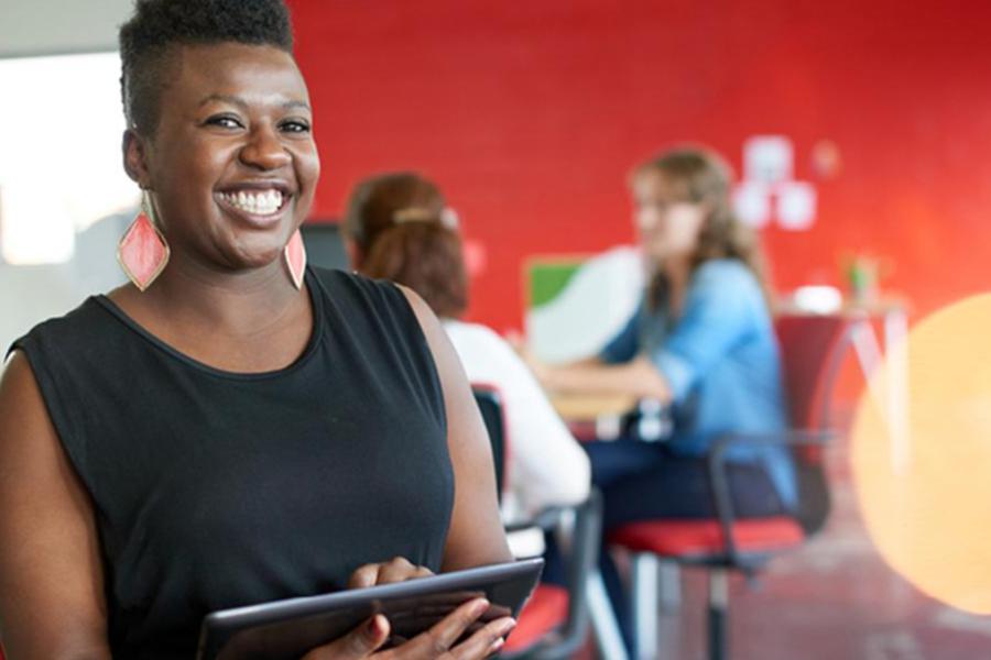 A smiling woman in a black top uses a tablet in a bright office with colleagues in the background.