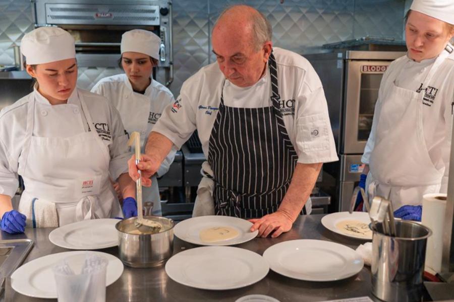 An experienced chef in a striped apron demonstrates plating a creamy sauce with a ladle while three culinary students in white uniforms and blue gloves observe closely in a professional kitchen.