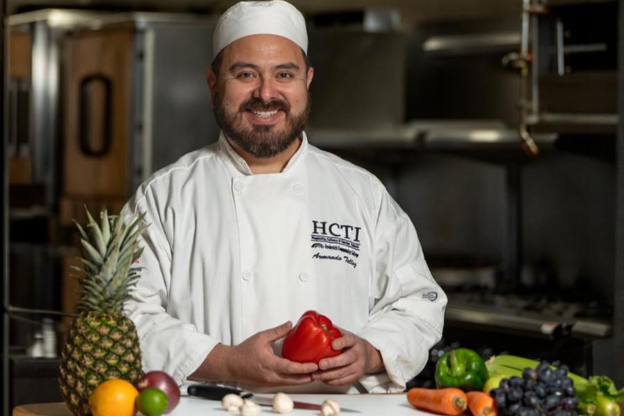 A smiling professional chef in a white uniform holding a red bell pepper with fresh produce in a kitchen.