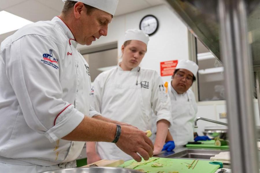 A professional chef instructor demonstrating knife skills to culinary students in a commercial training kitchen.