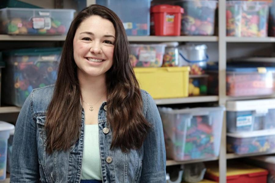 A smiling female educator or volunteer in a denim jacket standing in front of shelves organized with toys and supplies.