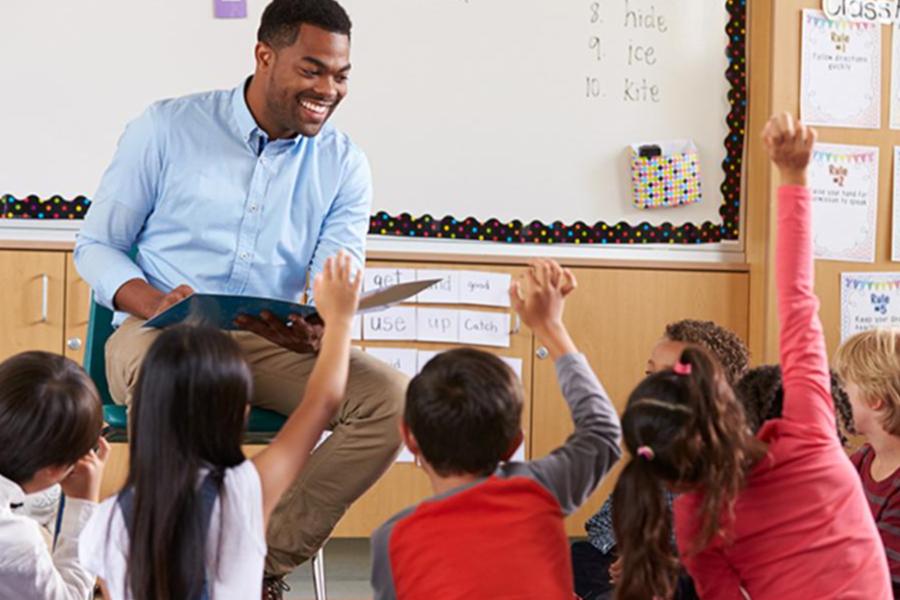 A smiling teacher reads to a group of young students sitting on the floor as they eagerly raise their hands to participate in the classroom.