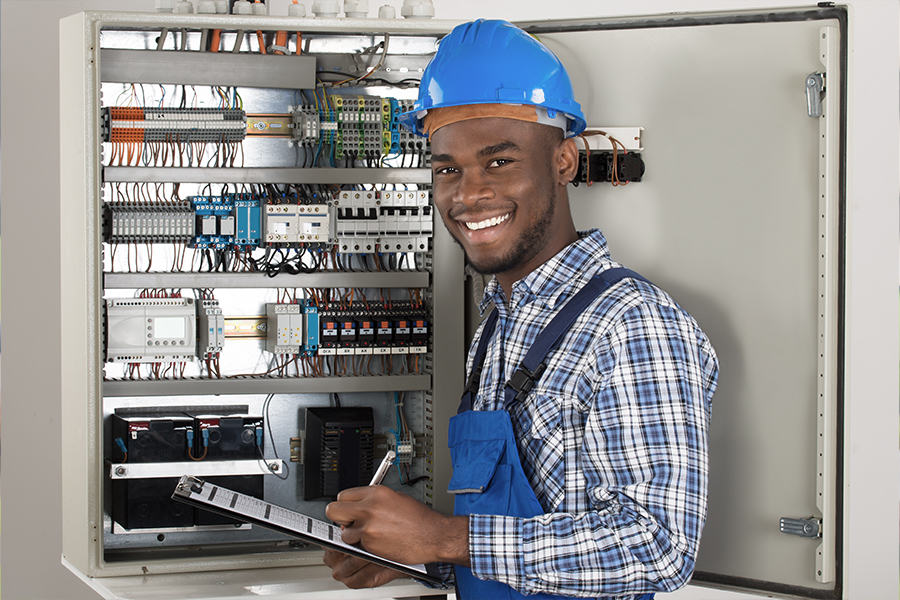 A smiling young electrician apprentice wearing a blue hard hat and plaid shirt holds a clipboard while standing in front of an open electrical panel.