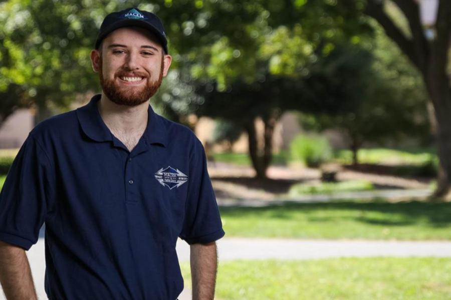 A smiling young man with a red beard wearing a navy blue polo and matching cap, standing in a sunlit park with trees in the background.
