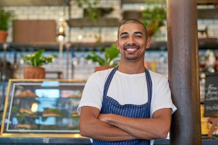 A smiling male cafe worker in a striped apron standing with arms crossed in a modern coffee shop.