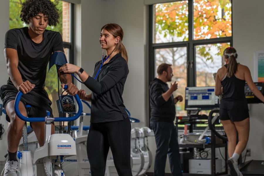 A student in a kinesiology lab uses a stethoscope and blood pressure cuff to monitor a male athlete on a stationary exercise bike.